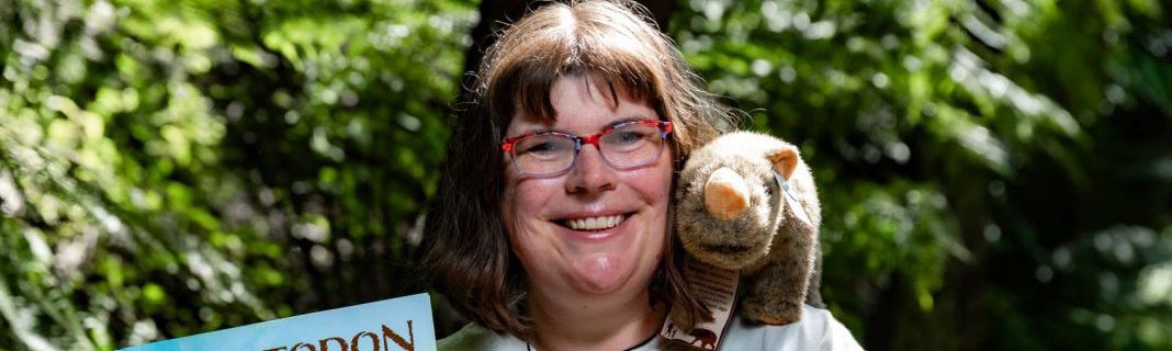 Bronwyn Saunders in a white shirt with a stuffed toy Diprotodon on her shoulder, holding a book about Diprotodons