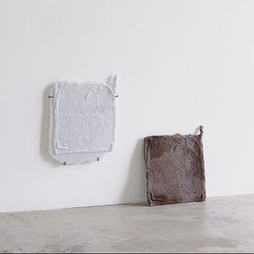 2 white and brown fabric squares leaning against a white wall, sitting on a polished concrete floor in a brightly lit room