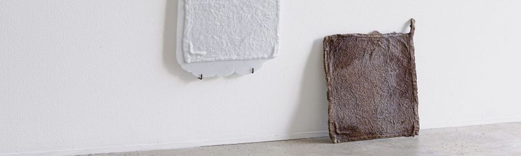 2 white and brown fabric squares leaning against a white wall, sitting on a polished concrete floor in a brightly lit room
