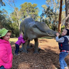 2 small children in cold weather clothes hugging a statue of a 4-legged megafauna creature with a crocodile-like mouth outdoors in front of gumtrees. In front of them stands a small girl in a pink jacket and green bucket hat. 
