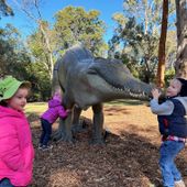 2 small children in cold weather clothes hugging a statue of a 4-legged megafauna creature with a crocodile-like mouth outdoors in front of gumtrees. In front of them stands a small girl in a pink jacket and green bucket hat. 