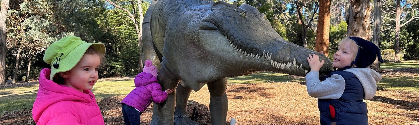 2 small children in cold weather clothes hugging a statue of a 4-legged megafauna creature with a crocodile-like mouth outdoors in front of gumtrees. In front of them stands a small girl in a pink jacket and green bucket hat. 