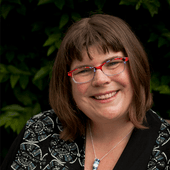 Jenny Dettrick smiling in red glasses and a blazer wearing a necklace, in front of a leafy backdrop